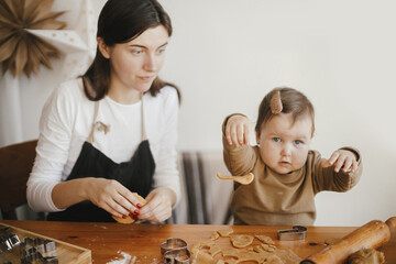 Adorable baby daughter together with mother making gingerbread cookies on wooden table in modern decorated scandinavian room. Cute funny toddler girl with christmas cookies. Mommy daughter moments
