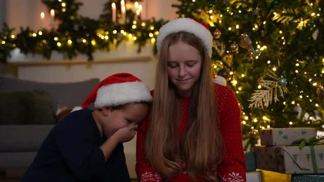 Joyful little brother and his older teenage sister wearing Santa Claus hats sitting on floor near fancy Christmas tree and laughing while watching funny video on tablet, family holiday leisure concept