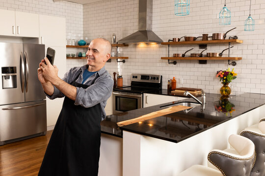 Man in kitchen wearing apron having a video call on his phone in kitchen. 