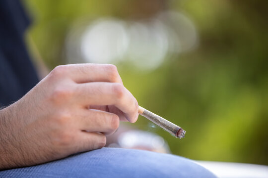 Detail of man holding a lit CBD pre-roll joint, cigarette with smoke rising sitting outdoors
