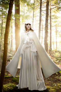Female ghost with black circles around her eyes, dressed all in white standing in a forest at sunrise