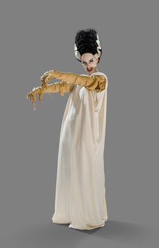 Full length portrait of a woman dressed up as the Bride Of Frankenstein, holding her arms out looking into camera smiling, against gray background