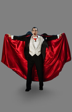 Portrait of a vampire against a gray background holding his cape out hissing, looking into camera