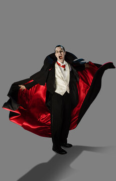 A vampire against a gray background holding his arms out at camera with mouth wide open