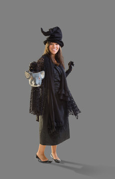 Full length shot of a Hispanic woman dressed as a witch for Halloween holding a mouse head trick or treat bucket dancing, against a white background