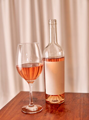 bottle of rosé wine with blank label with glass on wooden table with white curtain in the background. vertical. pink wine tasting