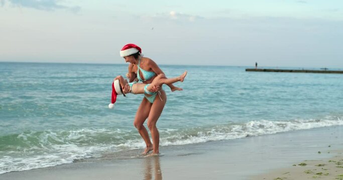 An adult woman in a swimsuit and a Santa hat catches a child running towards her with her hands and twirls around herself near the ocean.