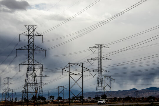 Huge Pylons And Electric Poles And Lines In Front Of A Car Junkyard Blurred Down Underneath With Stormy Sky And Mountains In Background And SUV Driving Down Road