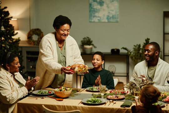 Portrait of caring African-American grandmother bringing food to table while celebrating Thanksgiving with big happy family
