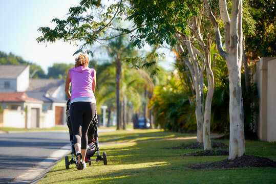 Back view of young mother pushing baby in stroller along urban street