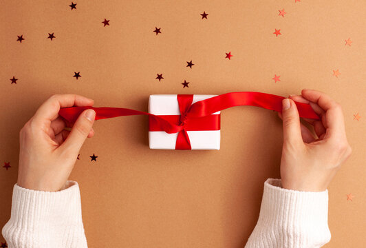 Human Hands In White Sweater Untied A White Paper Gift With A Red Satin Ribbon Bow On Brown Background With Red Stars Shapes Christmas Holidays Concept Flat La