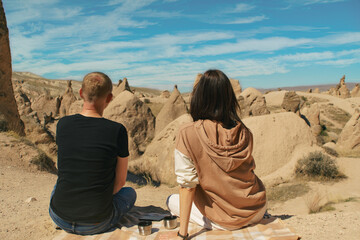 Couple of man and woman traveler sit on plaid and looking on landscape of spectacular volcanic rock formations in Cappadocia, Turkey