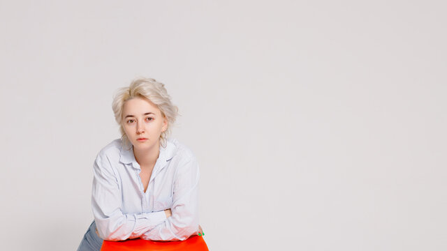 A blonde girl seriously looks at the camera, leaning her elbows on a chair on a gray background