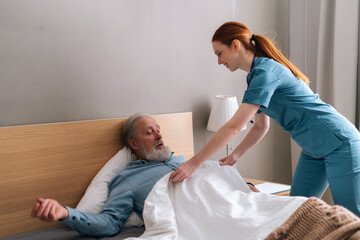 Cheerful young female nurse helping senior adult male patient to lying on bed and covering him blanket at home. Friendly woman taking care of mature old patient and talking to him