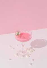 Cocktail glass with pink cocktail and silver christmas bauble in it on pastel pink and white background with fluorescent pearls. Christmas and new year party concept. Close up perspective.