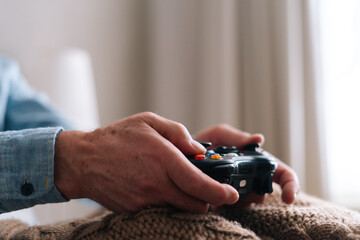 Close-up hands of unrecognizable senior male gamer playing online video game using gamepad at home. Closeup side view of mature elderly man playing computer game having fun with joysticks in bedroom. © dikushin