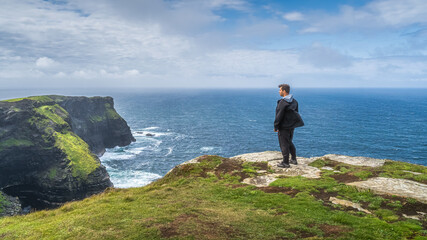 Man standing on the top of iconic Cliffs of Moher and admiring spectacular view, popular tourist...