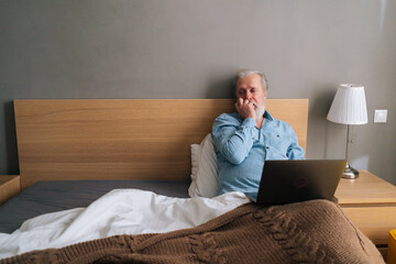 Thoughtful gray-haired senior male with beard using laptop computer lying sitting on comfortable bed at home. Handsome bearded mature older man in pajamas having online video call via notebook.