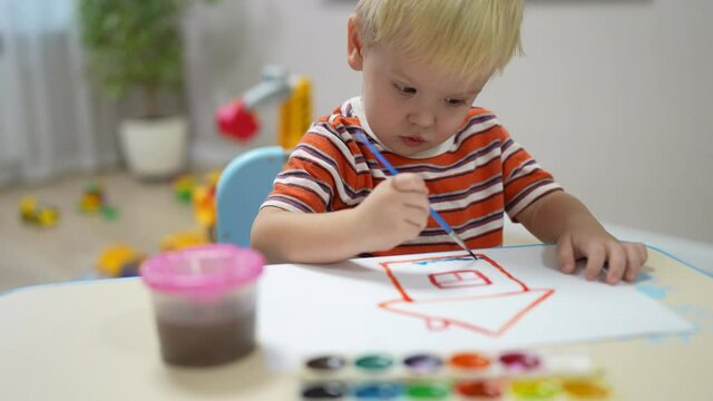 little boy with blond hair sits at the table and paints with paints on paper house.