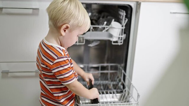 little boy with blond hair sits helping mom get the dishes out of the dishwasher