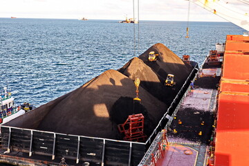 Loading coal from cargo barges onto a bulk carrier using ship cranes and grabs at the port of Muara...