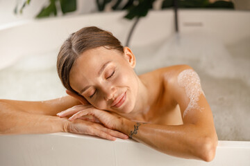 Portrait of peaceful young woman feeling relaxed, leaning on bathtub side, smiling with eyes closed while bathing. Wellness, beauty and care concept