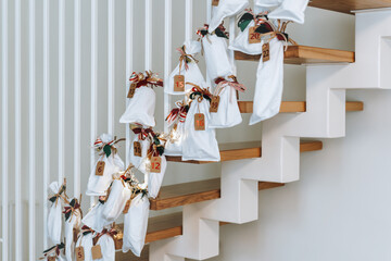 Advent calendar. White surprise bags decorated with colorful red-green Christmas ribbons. Handmade decorations hanging on stair railings in living room.