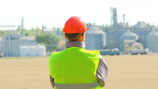 Grain elevator on farm. Engineer of maintenance in orange hardhat and yellow vest looks at built rural complex on autumn day backside view