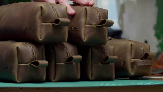 Camera moving along desk surface with many leather toiletry bags with different embossing names, craftsman hand picking up leather dopp kit indoors. Work process in handmade workshop