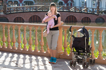 Mom with a baby girl and a stroller on the background of a pond and a bridge