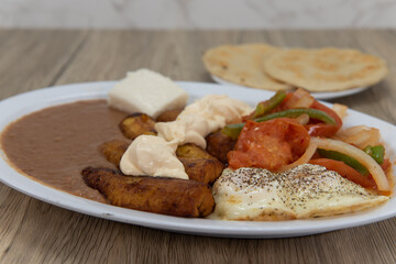 Fried plantains on a plate with refried beans, stir fried vegetables, and eggs served sunny side up with pupusa bread for a Latino meal
