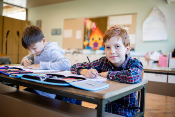 Portrait of concentrated schoolboy sitting at desk and writing in exercise book with classmate...