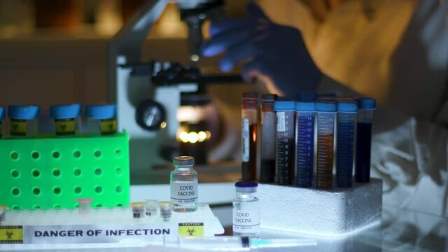 A medical laboratory technician or virologist using a microscope with COVID 19 vaccine vial in foreground.