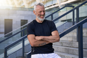 Joyful mature man in perfect shape smiling with eyes closed while standing with arms crossed after morning workout in the city