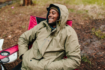 A young man sitting by the camp in a rain jacket / smiling. 