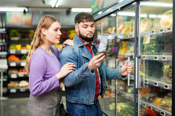 Young cheerful positive couple shopping together in grocery store, choosing packaged fresh greens...