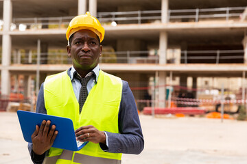 Portrait of an african-american engineer standing on the construction site, holding a laptop