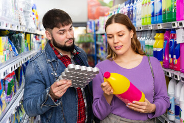 Young positive couple shopping at supermarket choosing detergent products
