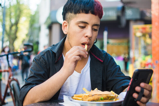 Young latino man eating fries while checking his phone - Powered by Adobe
