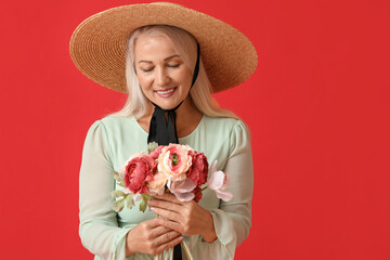 Beautiful mature woman in straw hat holding bouquet of flowers on red background