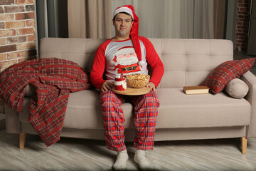 a young guy in a Santa Claus hat and pajamas is sitting on the couch holding a tray with popcorn and milk