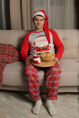 a young guy in a Santa Claus hat and pajamas is sitting on the couch holding a tray with popcorn...