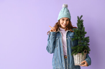 Stylish young woman in winter clothes and with Christmas tree showing something on color background