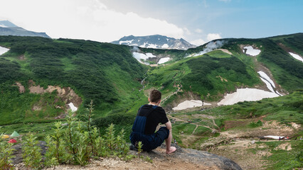 A man sits on a rock and looks down. There are tourist tents in the valley, people are walking along the paths. There is green vegetation and snow on the mountains. Steam from hot springs is visible.