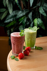 Different smoothies in misted cold glasses, standing on wooden round table, top view, green foliage on blurred background