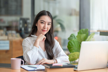 Successful smiling beautiful young Asian businesswoman sitting with laptop and computer while doing some paperwork at the office. Looking at camera.