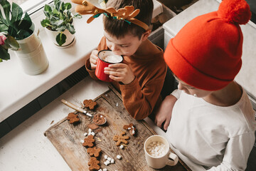 Children drink cocoa with marshmallows and christmas gingerbread man on a wooden background. Winter vacation at home. Fun time in the kitchen. 2 boys with antlers are cooking in the kitchen