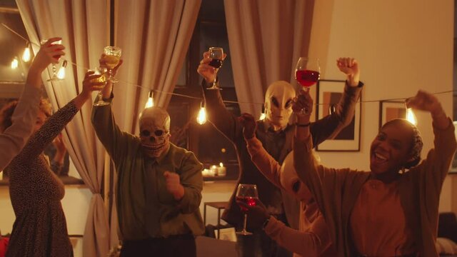 Slow motion shot of company of friends in scull and alien masks holding wine glasses and dancing together at Halloween party