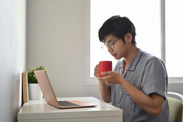 Peaceful asian man in pajamas using computer laptop in bedroom and drinking coffee.