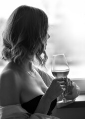 Beautiful young girl with a glass of wine sits near the window. Black and white photo.